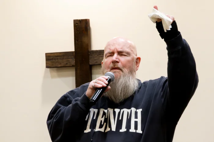 Worship leader singing and raising hand with cross in background.